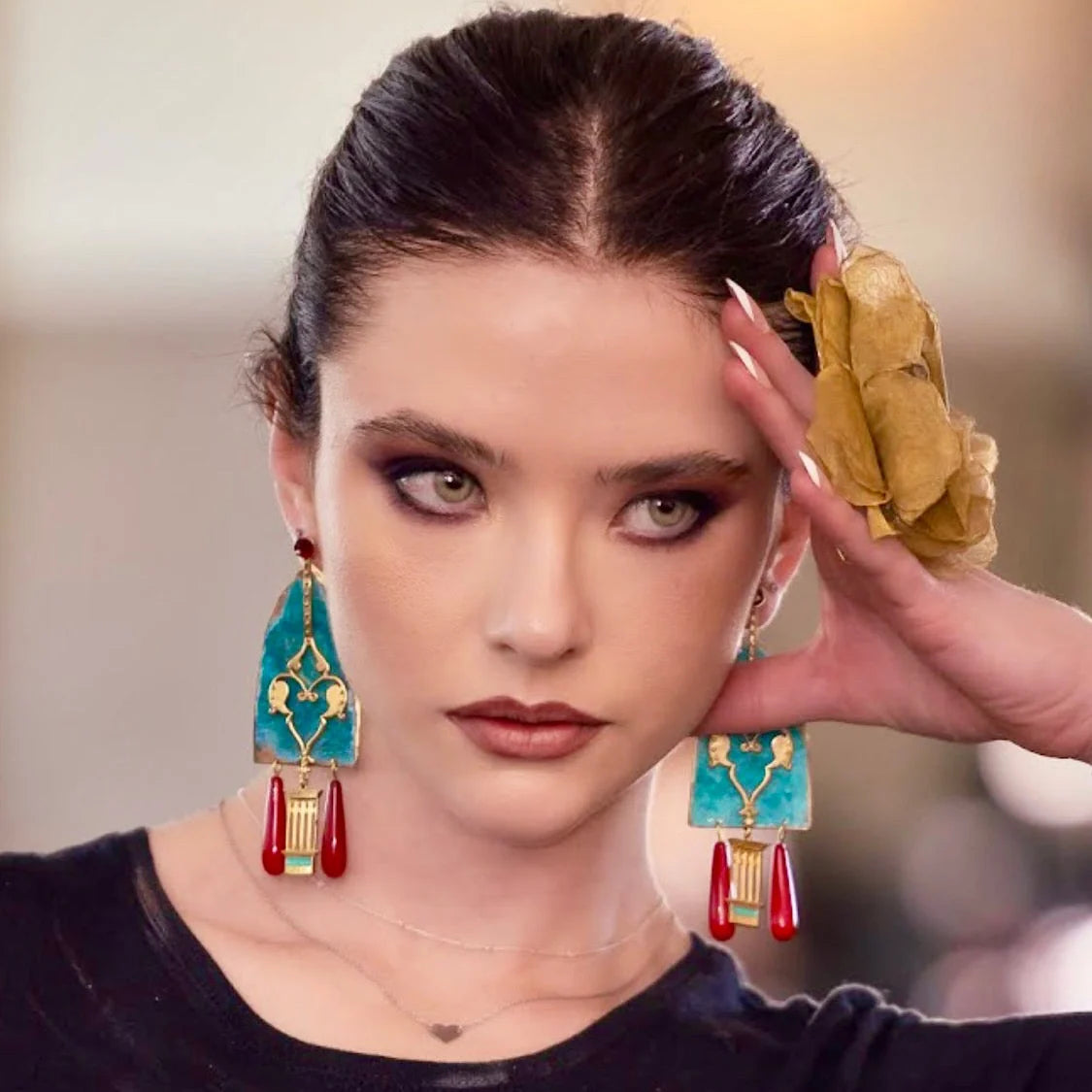 A close-up of a model wearing large turquoise and gold statement earrings with dangling red tassels, with her hair pulled back and a gold flower accessory in her hand.